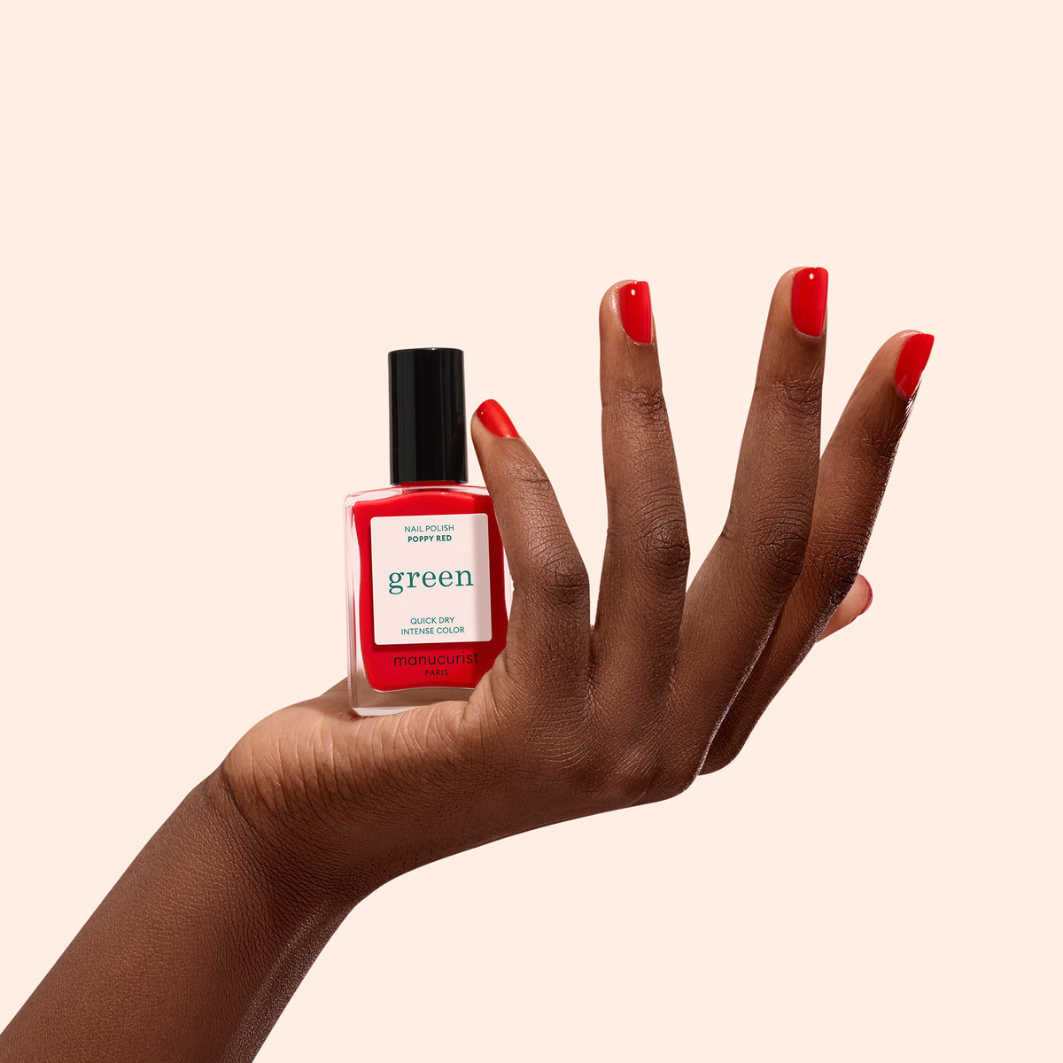 A hand with neatly painted Poppy Red nails holds a bottle of Manucurist Poppy Red nail polish against a plain light beige background.