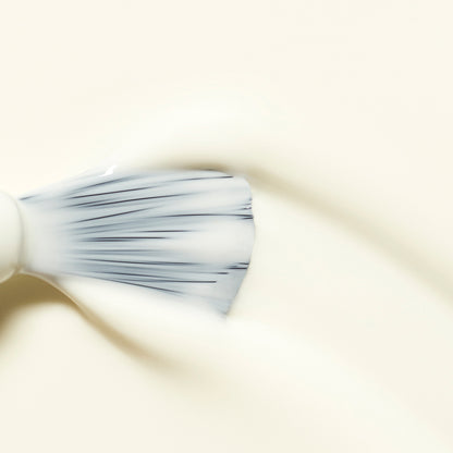 Gros plan sur un pinceau étalant la base traitante blanc crème Porcelaine de Manucurist sur une surface lisse et claire. Les poils sont écartés pour répartir uniformément le produit.