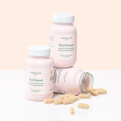 Three bottles of Manucurist Nail Boost supplements are arranged on a light surface, one lying on its side with beige capsules spilled out, highlighting support for nail growth and stronger nails.