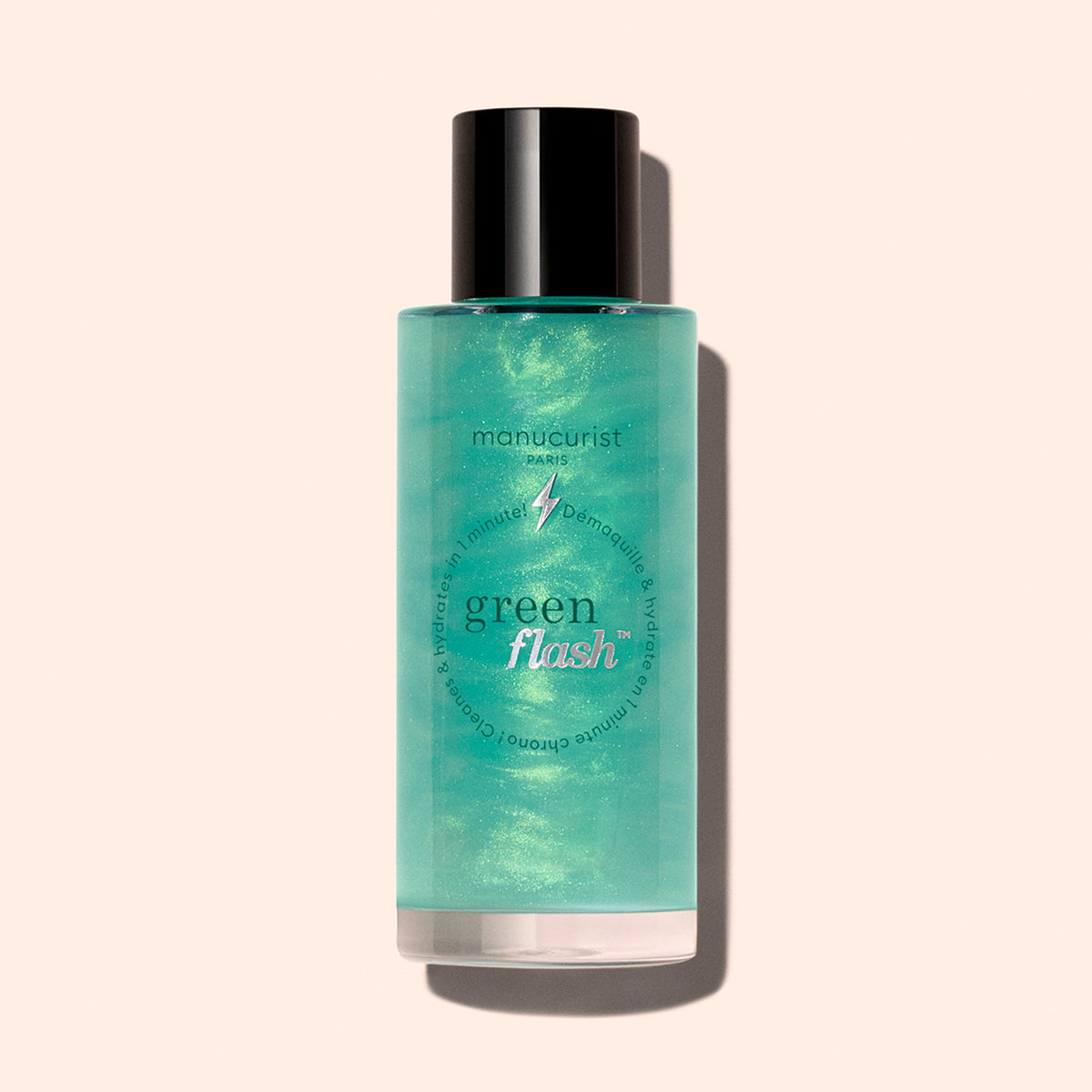 Un flacon de dissolvant doux pour paillettes Green Flash™ de Manucurist avec un bouchon noir, contenant un liquide vert clair enrichi en huile de ricin, est présenté sur un fond beige clair.