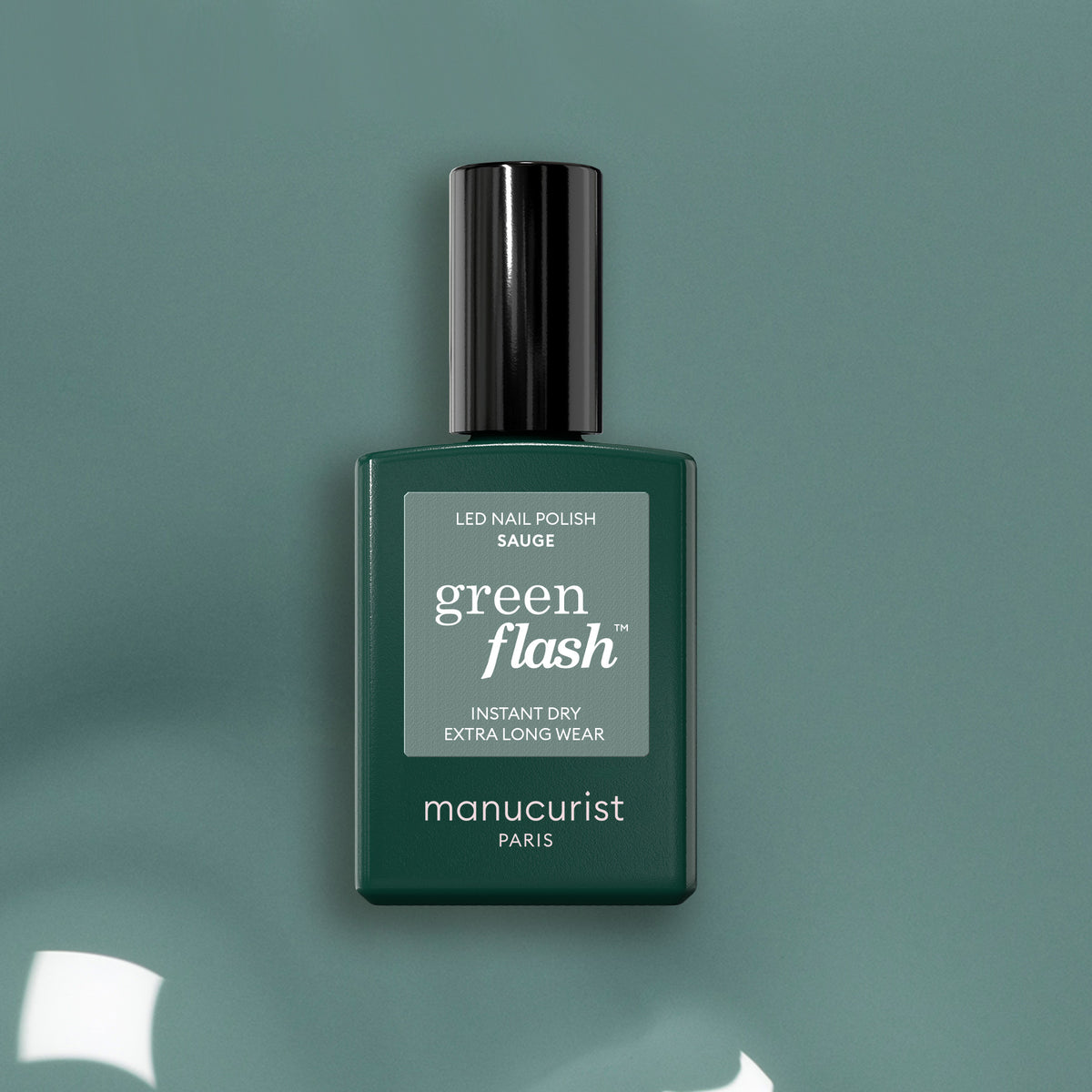 Un flacon de vernis à ongles LED Green Flash Sage de Manucurist se tient debout sur une surface vert sauge, avec un bouchon noir brillant et une étiquette minimaliste qui met en valeur sa douce teinte verte.