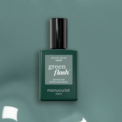 Un flacon de vernis à ongles LED Green Flash Sage de Manucurist se tient debout sur une surface vert sauge, avec un bouchon noir brillant et une étiquette minimaliste qui met en valeur sa douce teinte verte.