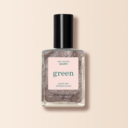 A clear bottle of Manucurist Quartz nail polish with a black cap, showcasing a shimmery light rose gold hue labeled “green” and “quick dry intense color” on a soft beige background.