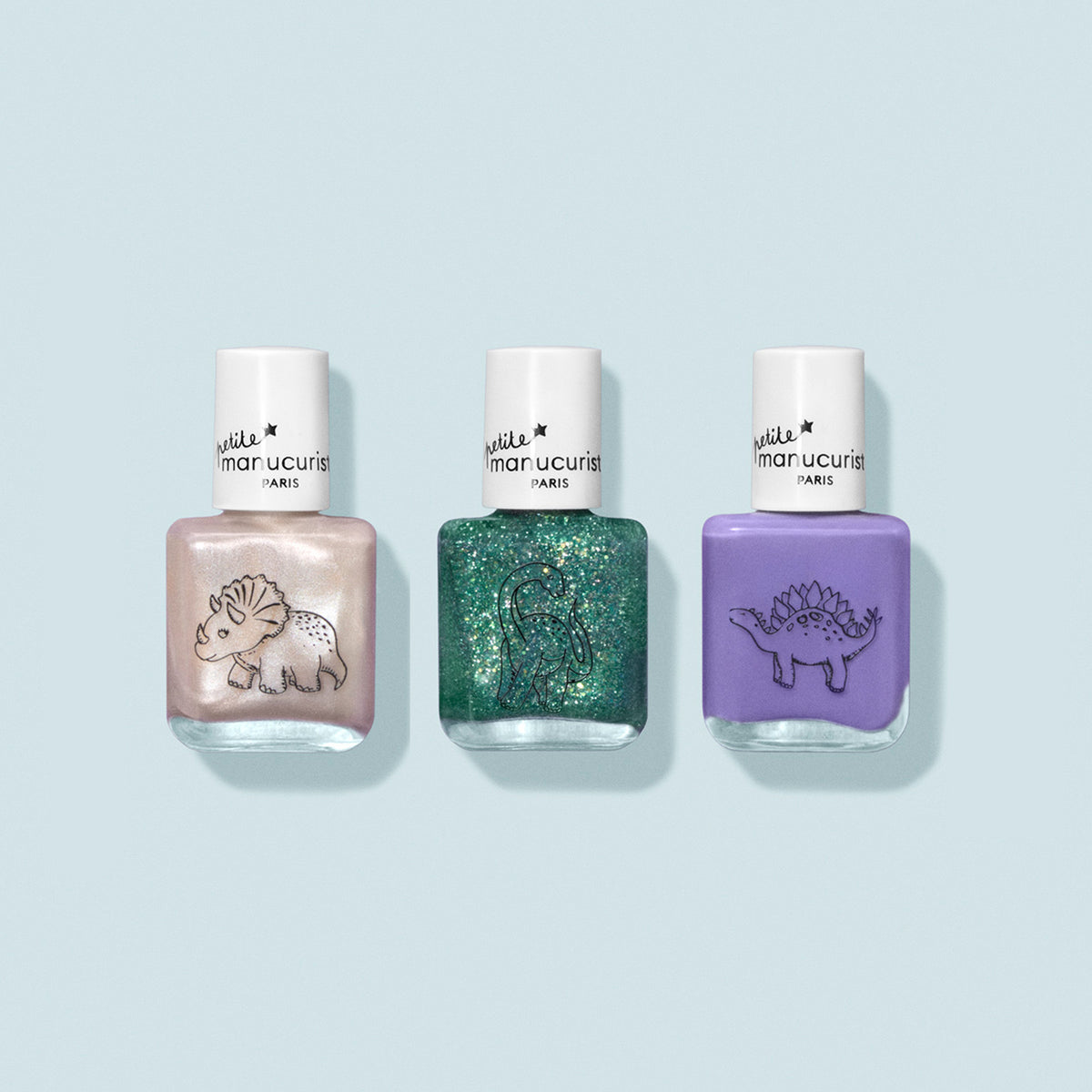 Le coffret de vernis à ongles pour enfants Wild Life Collection de Manucurist comprend trois flacons à la formule douce : blanc nacré avec un tricératops, vert pailleté avec un brontosaure et violet pastel avec un stégosaure, le tout sur fond bleu pâle.