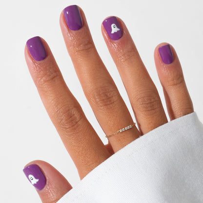 A hand sports Manucurist’s Witchy  Wicked purple gel polish, with two nails adorned with white ghost Halloween art. A thin gold ring is worn, and a white sleeve partially covers the hand.