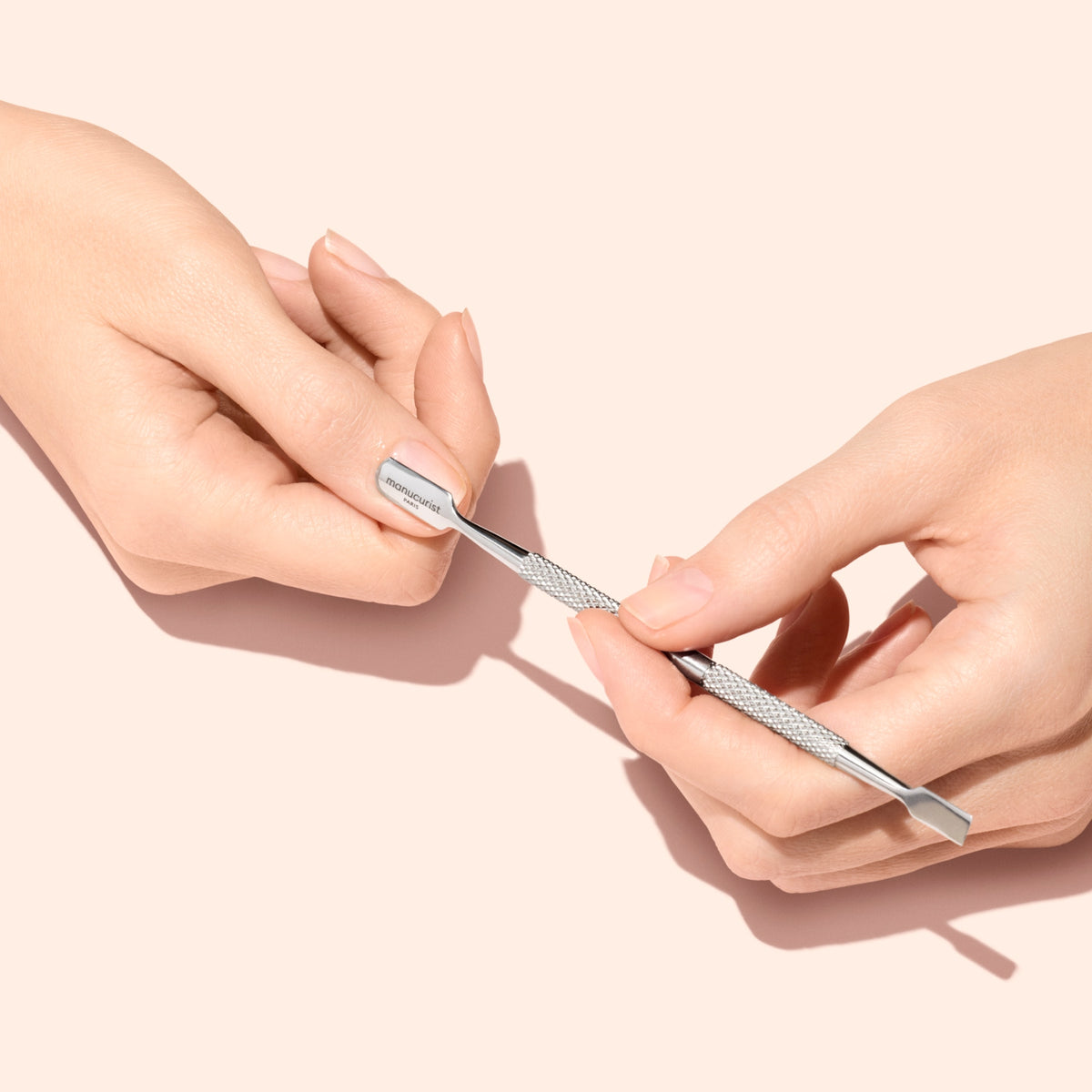 A person uses a metal cuticle pusher to gently push back cuticles on a fingernail. The background is a solid light pink color.