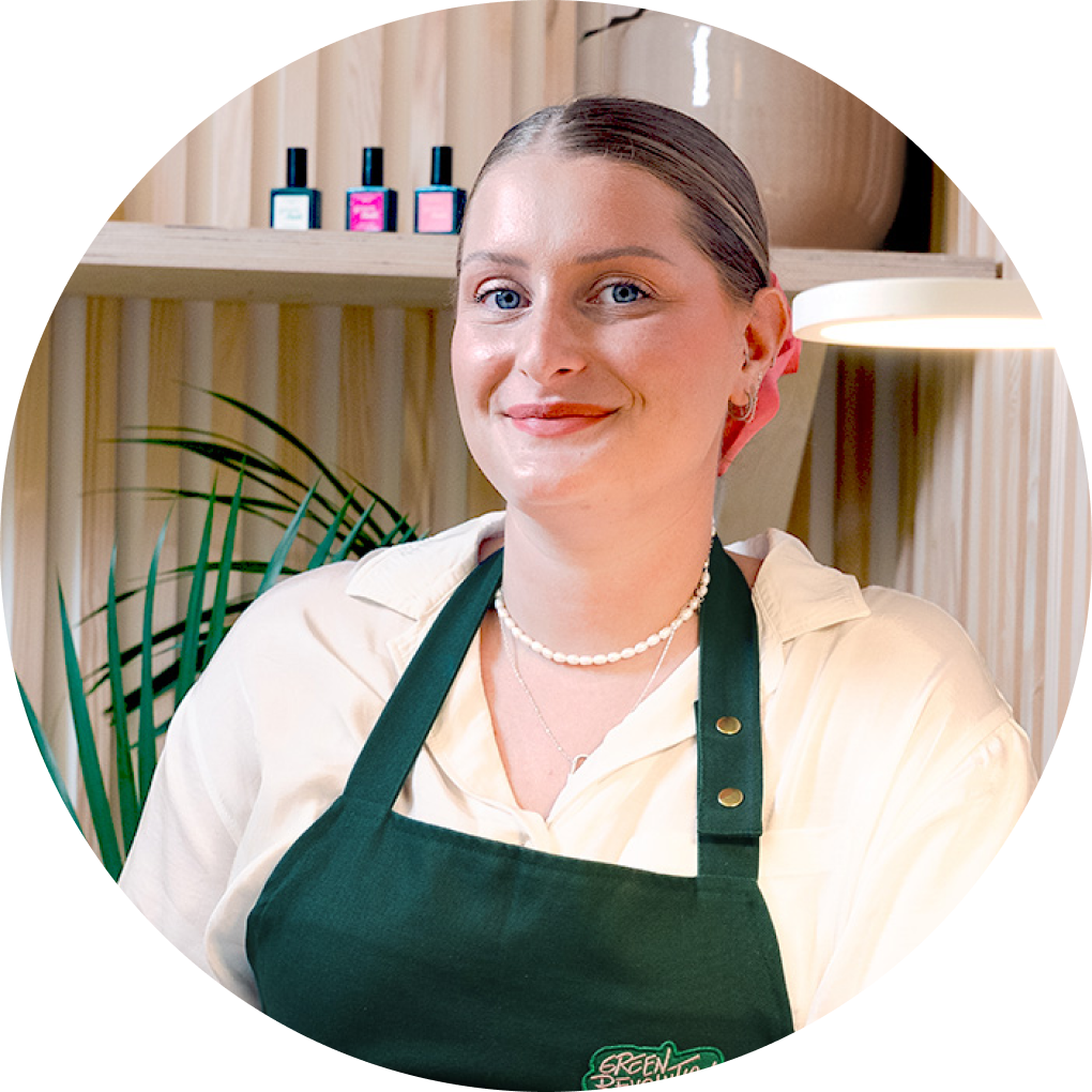 A woman with light hair tied back, wearing a white shirt, green apron, and pearl necklace smiles at the camera. She stands indoors with wooden shelves, green plants, and colorful bottles in the background.
