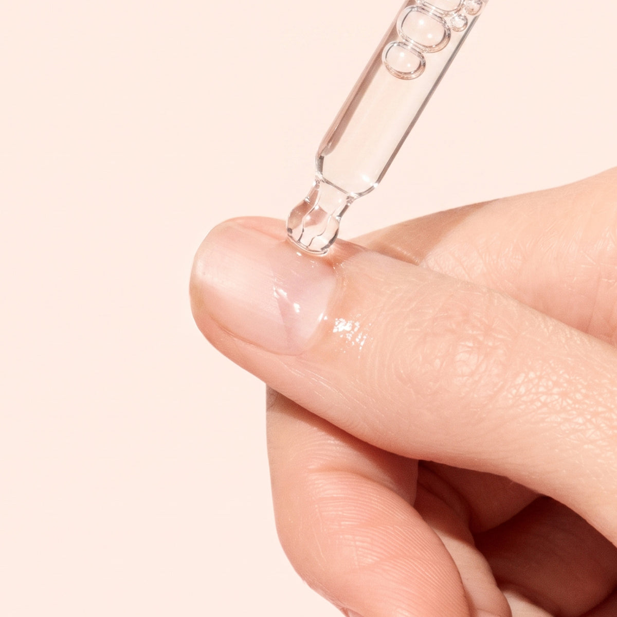 A close-up of a hand applying clear serum to a fingernail with a glass dropper against a light beige background.