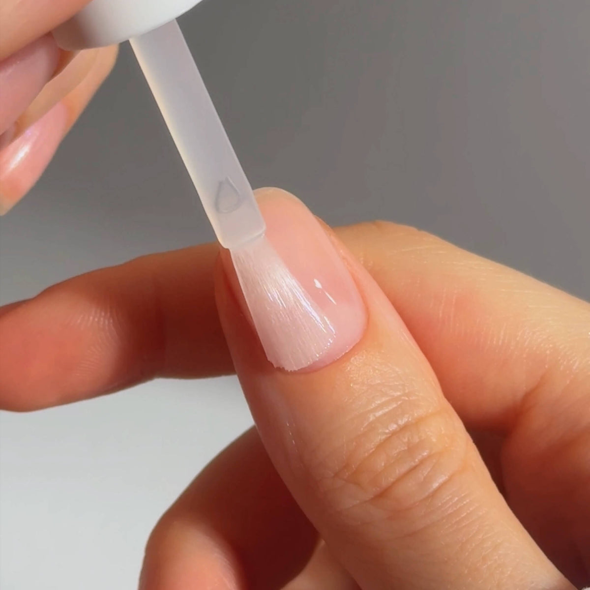 A hand applies clear nail polish to a fingernail using a brush applicator, with the nails neatly shaped and the background softly blurred.