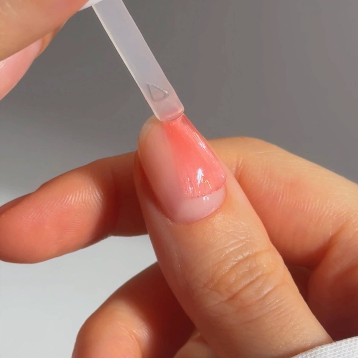 A close-up of a hand with a clear nail polish brush applying a layer of light pink polish to a fingernail. The nail is clean and well-shaped, and the background is softly blurred.