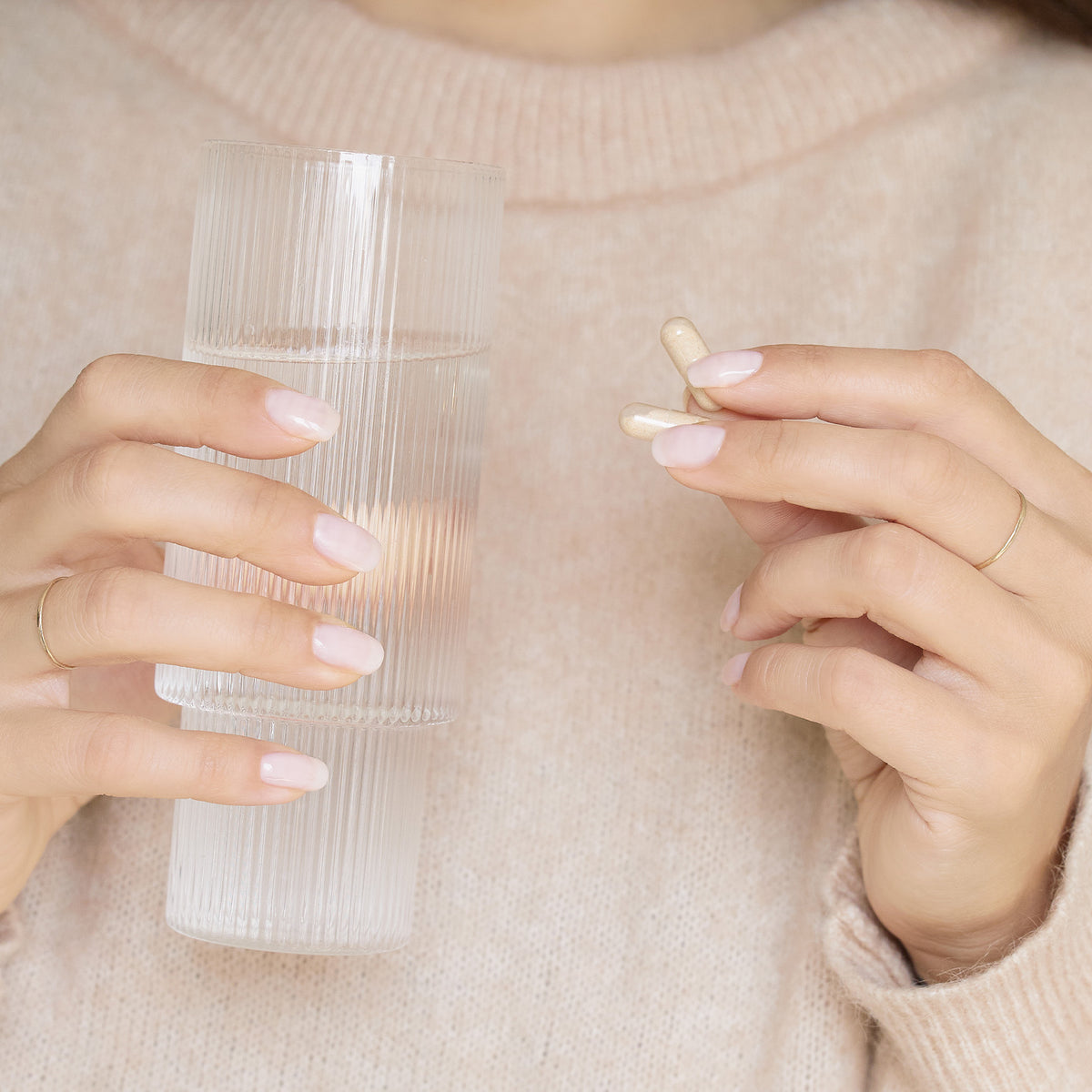 A person in a beige sweater holds a ribbed glass of water in one hand and a beige capsule pill in the other, with neatly manicured nails visible.