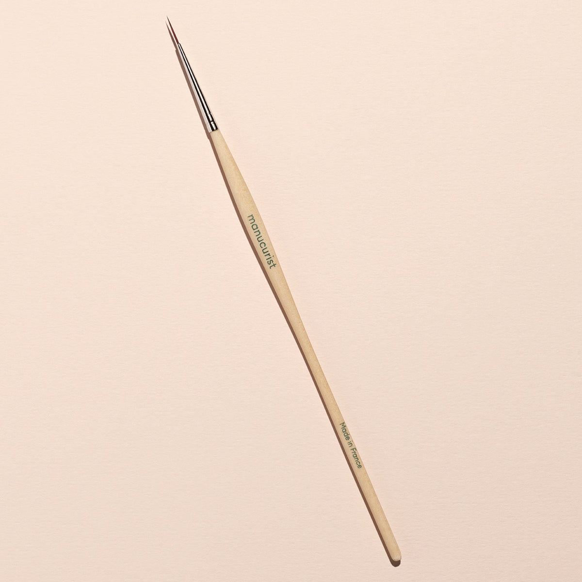 The Witchy  Wicked brush by Manucurist, featuring a wooden handle and metal ferrule, lies diagonally on a light beige surface—ideal for detailed Halloween nail art with gel polishes or adding biodegradable glitter accents.