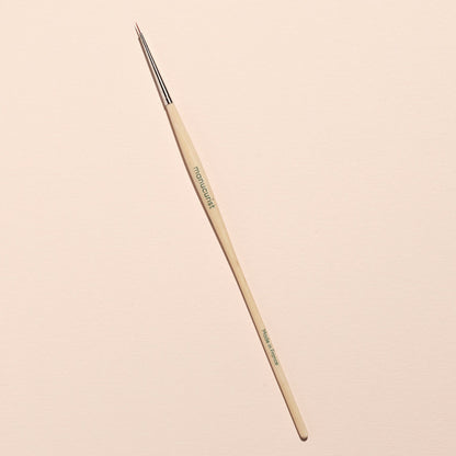 The Manucurist Pumpkin Patch paintbrush, featuring a thin wooden handle and fine pointed tip ideal for detailed Halloween nail art, is placed diagonally on a light beige background.