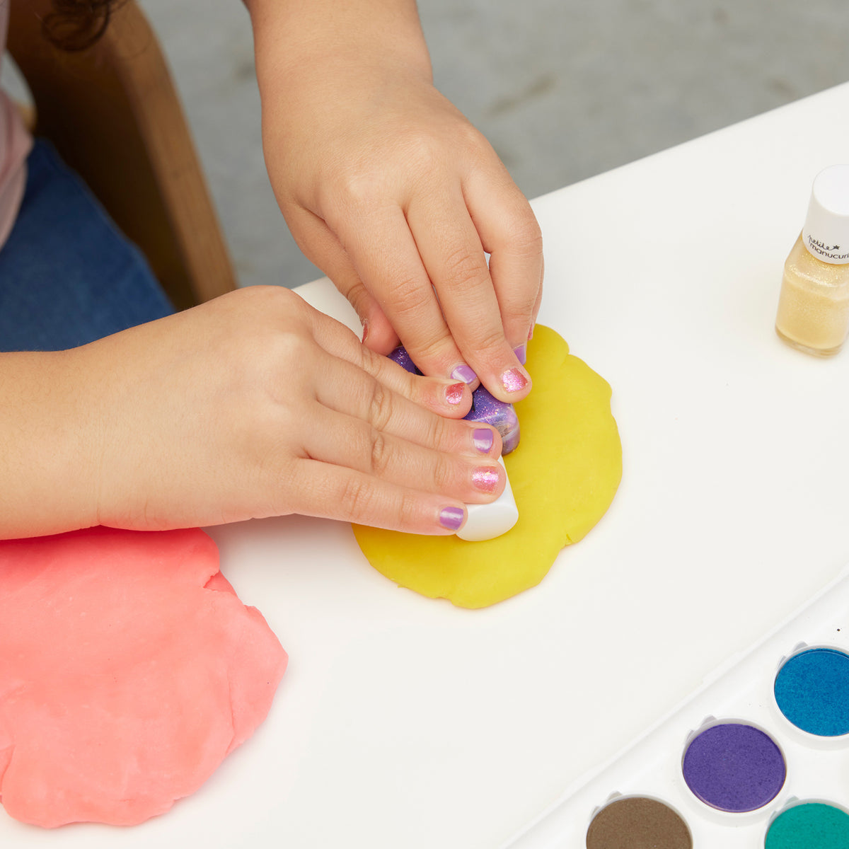 Un enfant portant du vernis à ongles coloré du Trio Manucurist Betty Scarlett Stella presse un tampon dans de la pâte à modeler jaune sur une table blanche, tandis que de la pâte à modeler rose et des peintures aquarelles sont éparpillées à proximité.