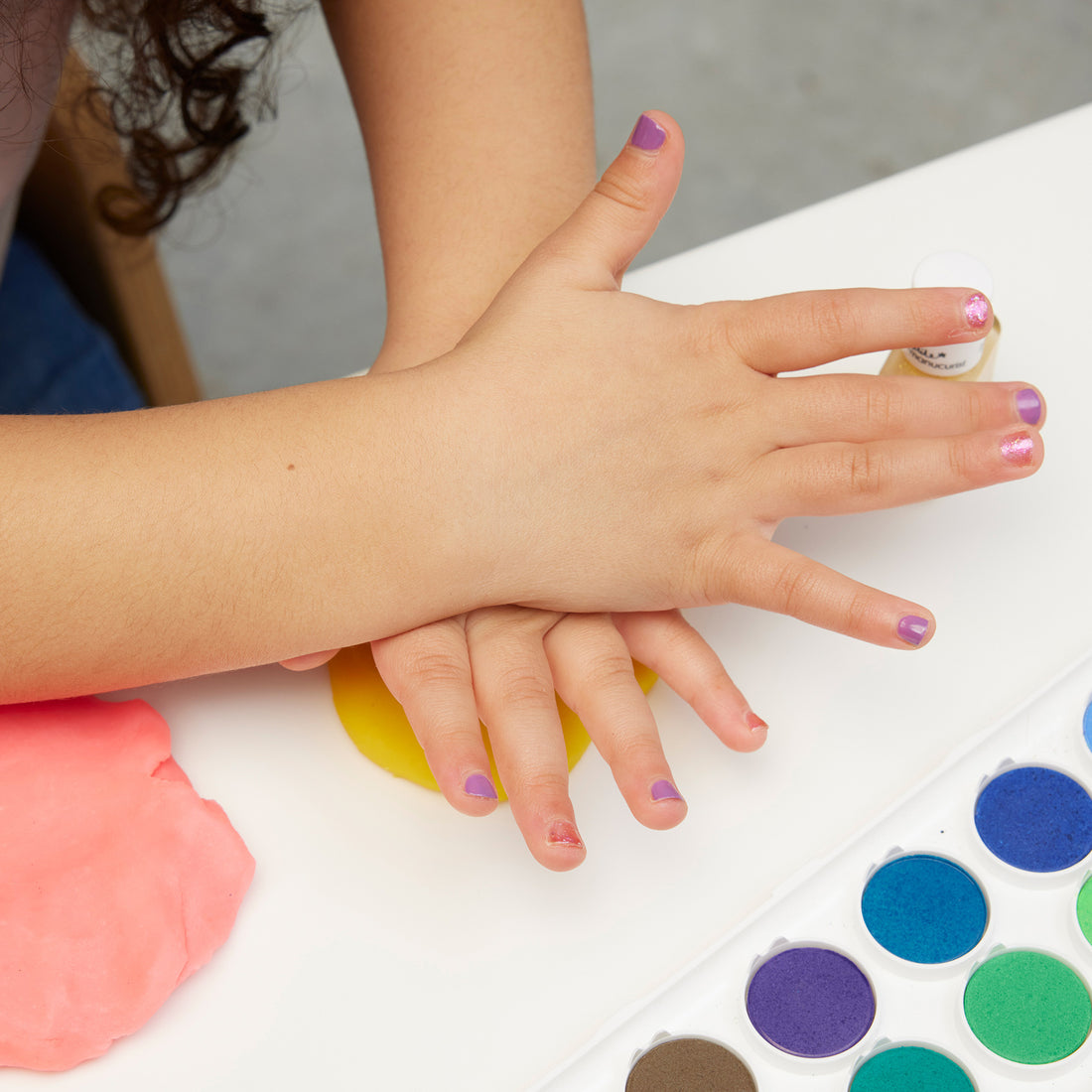 Un enfant avec le Trio de vernis à ongles non toxiques Bonnie Stella Janis de Manucurist presse ses mains sur de la pâte à modeler jaune et rose, à côté d’une palette d’aquarelle sur une table blanche.
