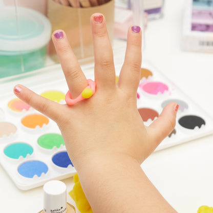 La main d’un enfant aux ongles colorés utilisant le Trio Betty Scarlett Stella de Manucurist est présentée au-dessus d’une palette de peintures aquarelle, de fournitures artistiques et d’une bague rose et jaune dans un espace de travail lumineux et créatif.