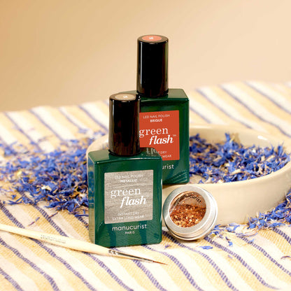 Two bottles of Manucurist Oriental Vibe nail polish and a pot of gold biodegradable glitter from the Autumn 2023 collection sit in a beige bowl on a striped cloth, surrounded by dried blue and purple flower petals. A manicure brush lies nearby.