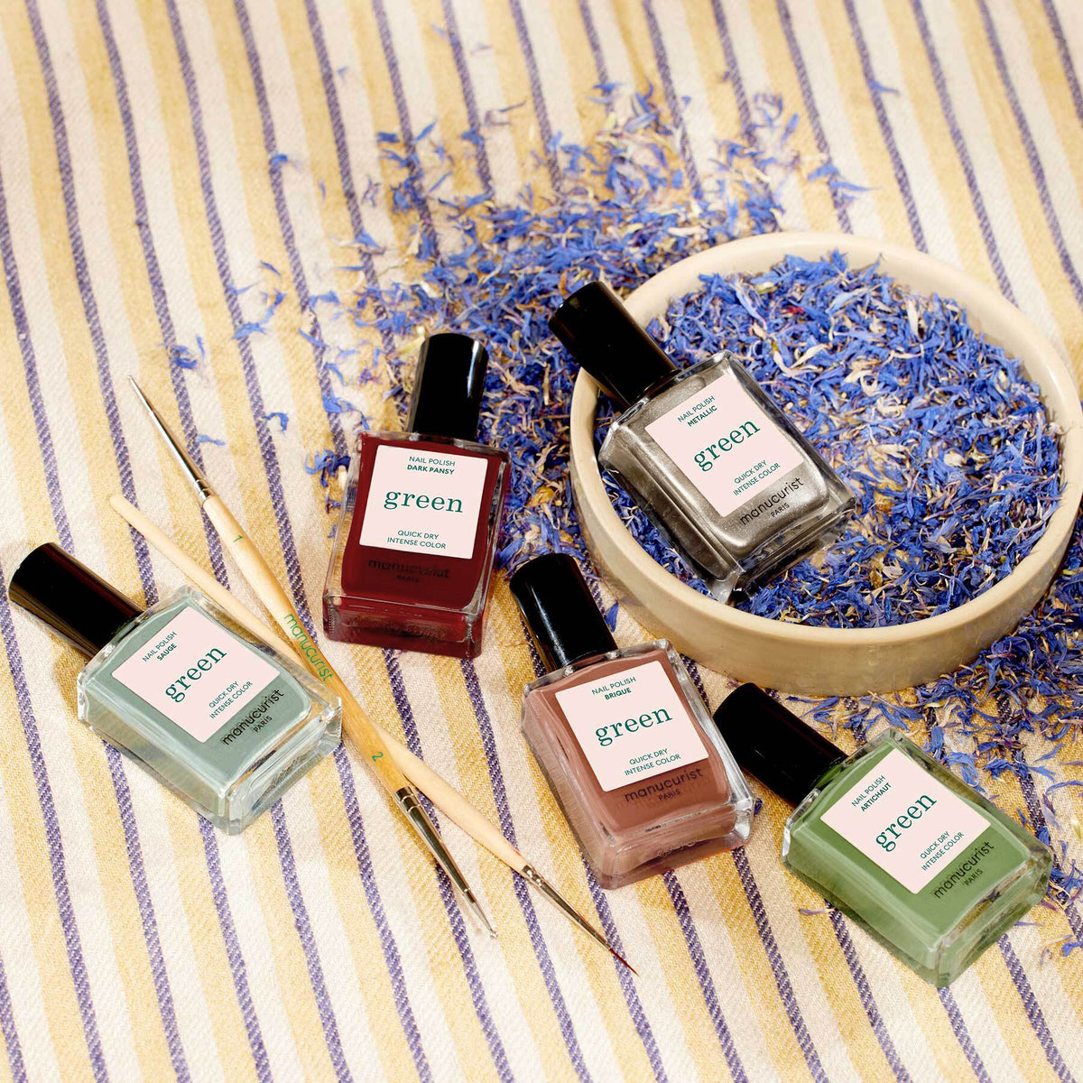 Five bottles of Manucurist’s Green Flash green nail polish from the Autumn 2023 collection are displayed on striped fabric with blue dried flowers, a wooden bowl, and two Manucurist Spice Lover nail art tools nearby.