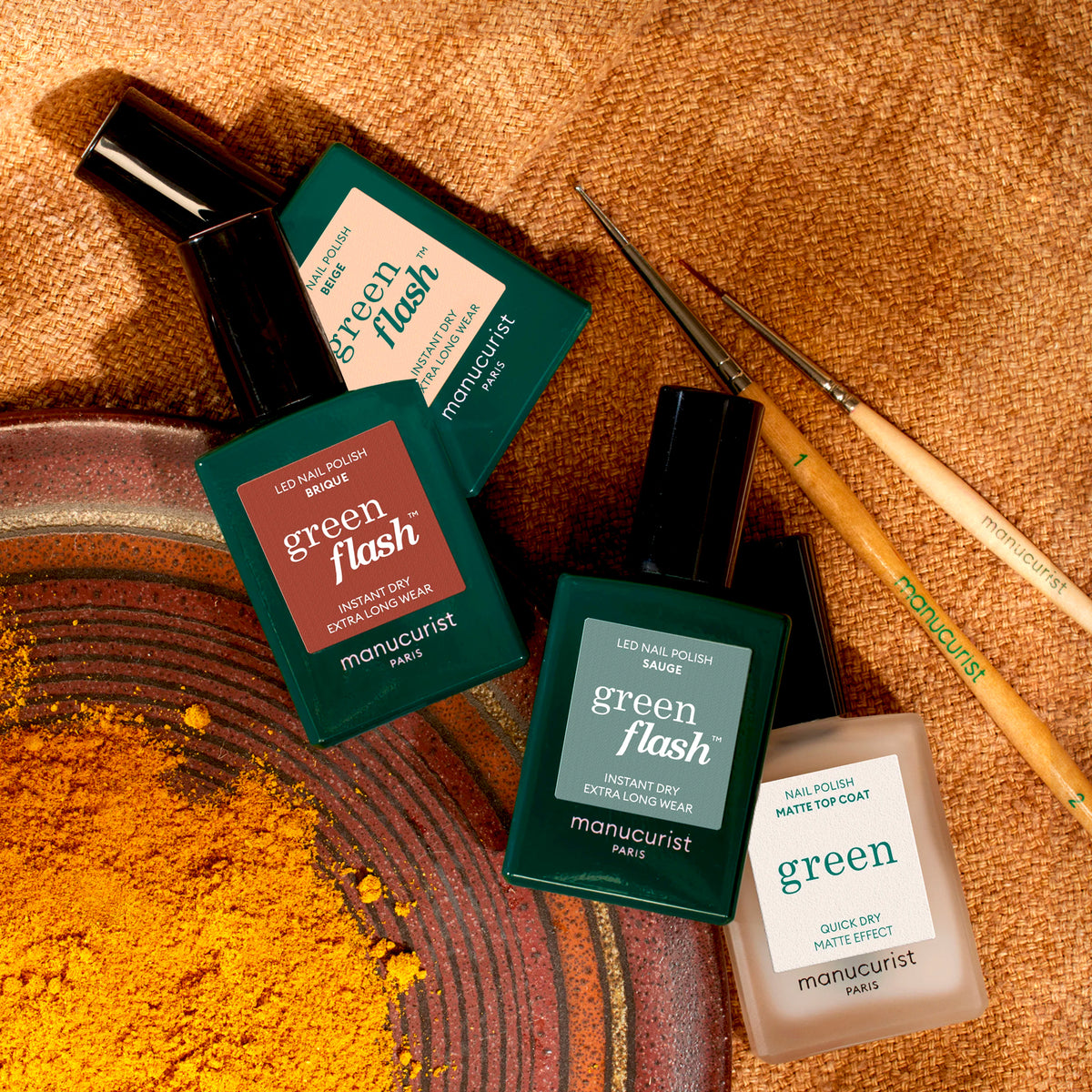 Four bottles of Manucurist Geometric Obsession nail polish in green and cream shades from the Autumn 2023 collection are displayed on a textured brown surface with yellow powder, two nail art brushes, and geometric nail art inspiration.