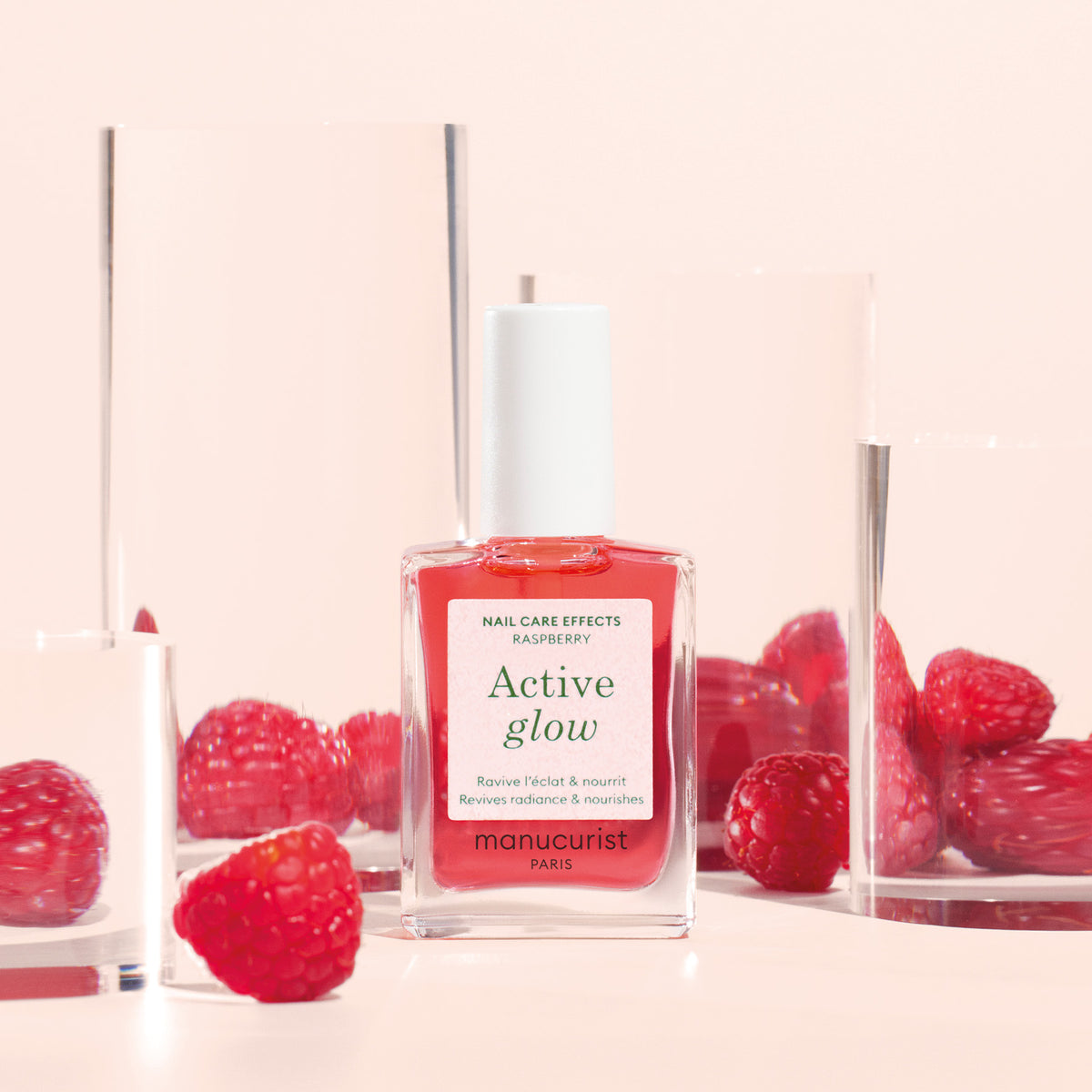 A bottle of Manucurist Active™ Glow Raspberry nail care rests among clear acrylic blocks and fresh raspberries on a pink background. The label emphasizes its plant-based formula, promoting radiant nails and nourishing care infused with the fruity benefits of raspberry.
