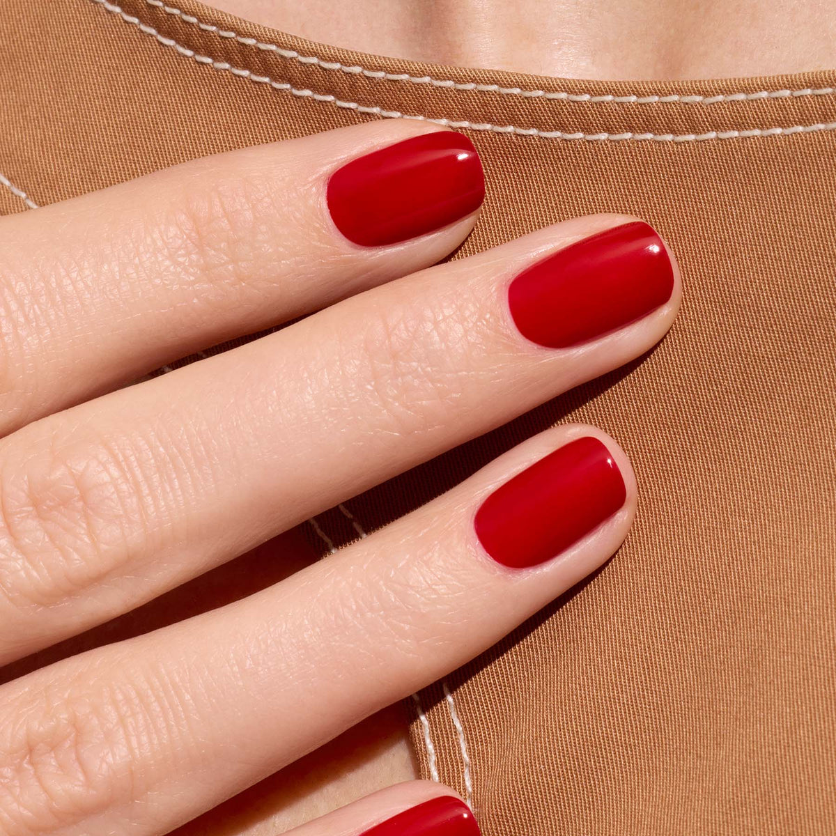 A close-up of a hand with neatly manicured nails painted in Manucurist’s Red Cherry polish, held against tan fabric with white stitching.