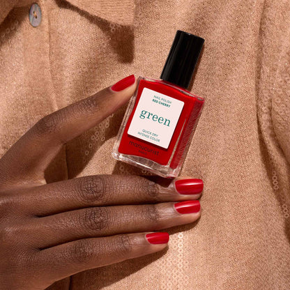 A hand with neatly manicured red nails holds a bottle of Manucurist Red Cherry nail polish against a beige, textured fabric background.