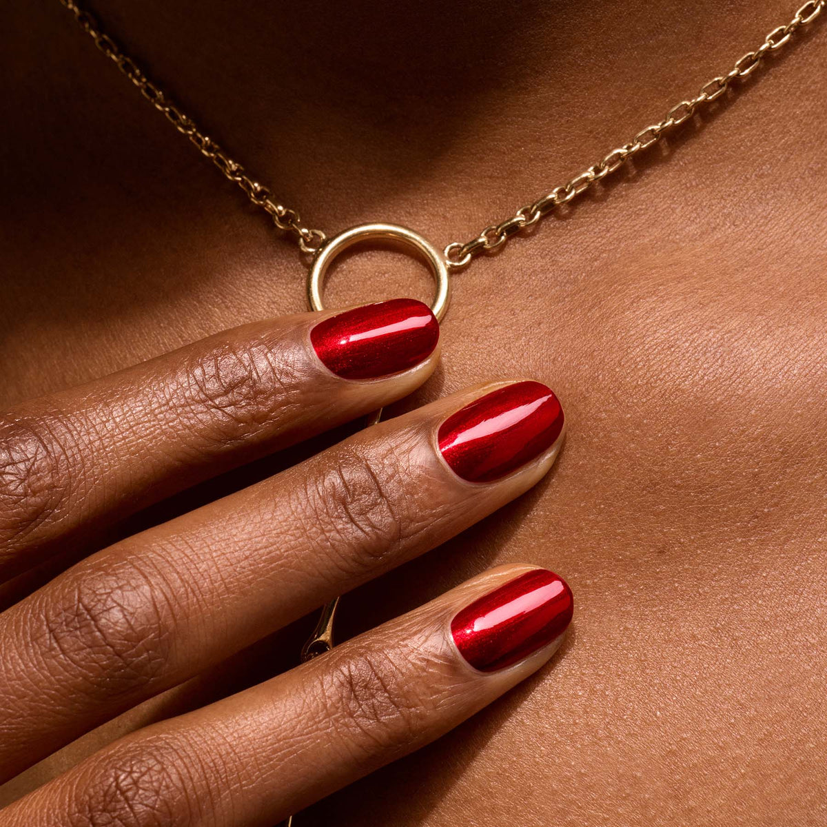 Gros plan sur une main portant le vernis Manucurist Red Hibiscus posée sur un décolleté, mettant en valeur un collier doré orné d&