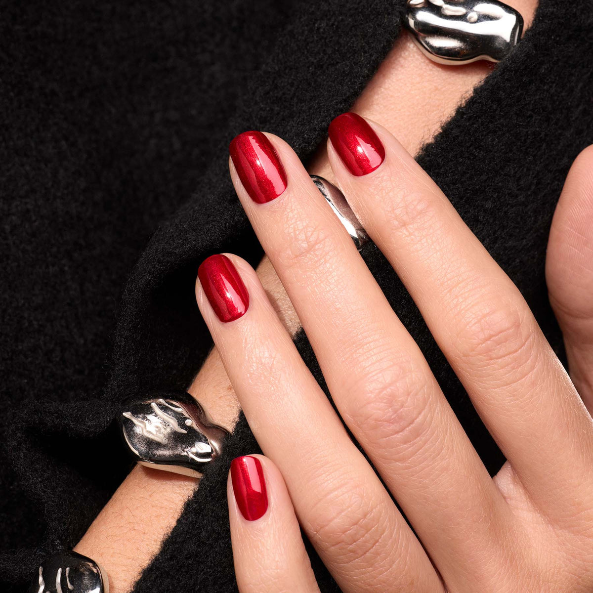 Gros plan d’une main avec le vernis Red Hibiscus de Manucurist, posée sur un tissu noir texturé. Le look est complété par d’épaisses bagues argentées et un bracelet assorti aux formes lisses et organiques.
