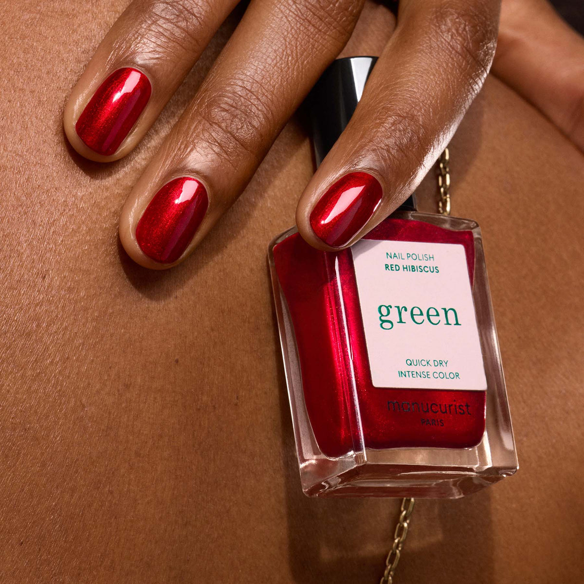 Une main aux ongles peints en rouge tient un flacon de vernis à ongles Manucurist Red Hibiscus sur une peau brune. Cette teinte vive et brillante ne passe pas inaperçue.