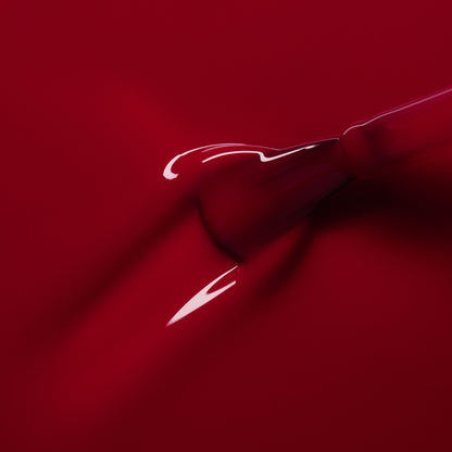 A close-up shows a dropper releasing Manucurist’s Pomegranate gel polish—a glossy raspberry red—onto a smooth red surface, forming a shiny, flowing puddle.