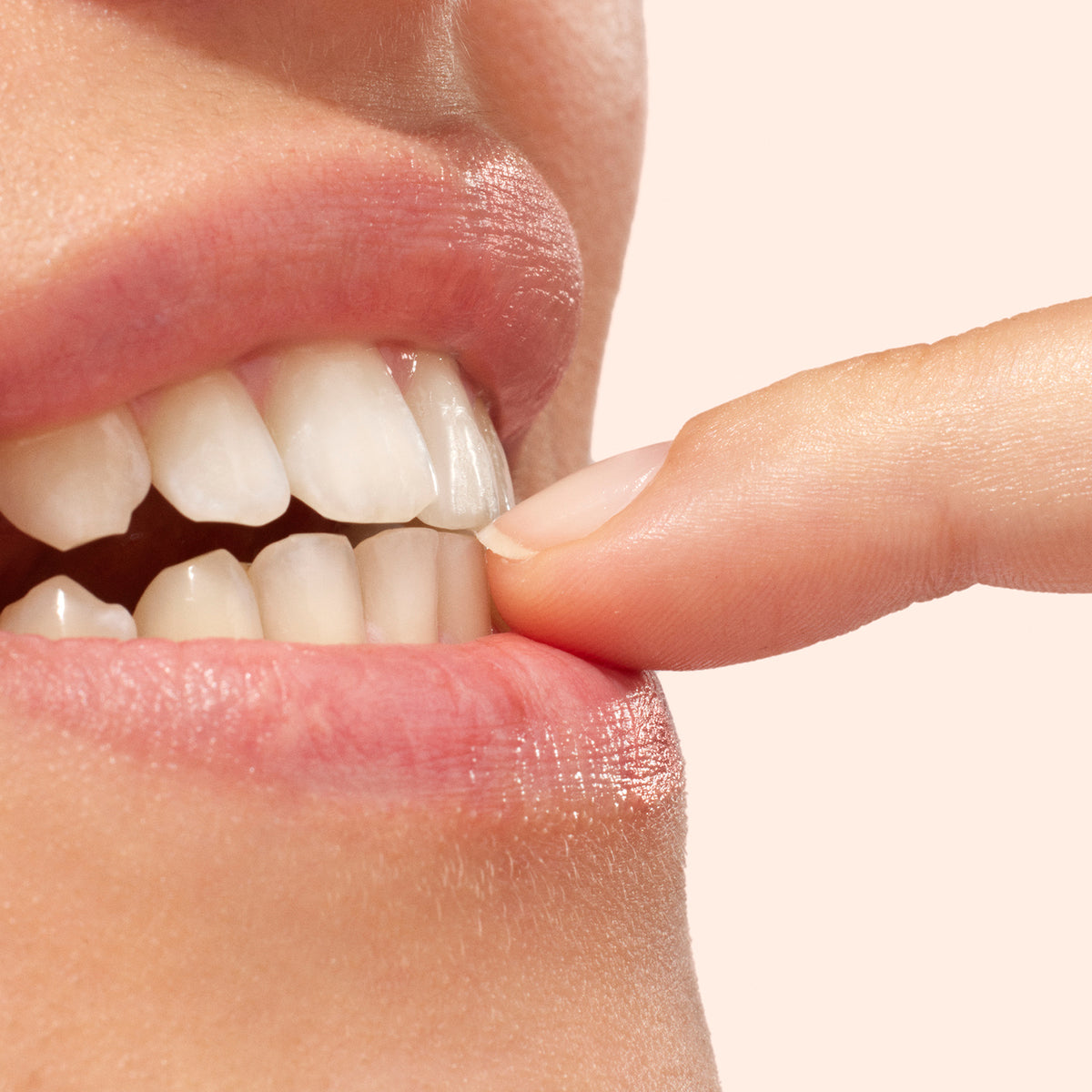 A close-up of a mouth with white teeth and pink lips, as the index finger—coated in Manucurist&
