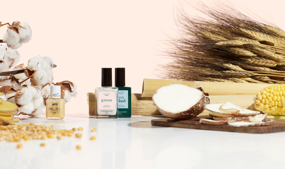 Bottles of nail polish are displayed on a white surface, surrounded by cotton, wheat, sugarcane, corn kernels, and sliced cassava on a wooden board, with a light pink background.