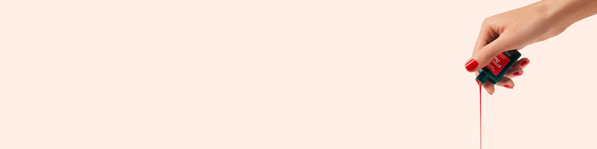 Une main aux ongles vernis de rouge incline un flacon de vernis à ongles rouge, laissant couler le vernis sur un fond beige clair. L’image est minimaliste, avec la majeure partie de l’espace vide, à l’exception de la main et du flacon à droite.