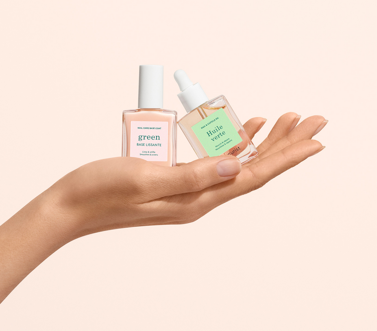 A hand holds two small glass bottles of nail care products with pastel labels on a light beige background; one is a nail base, and the other is cuticle oil with a dropper.