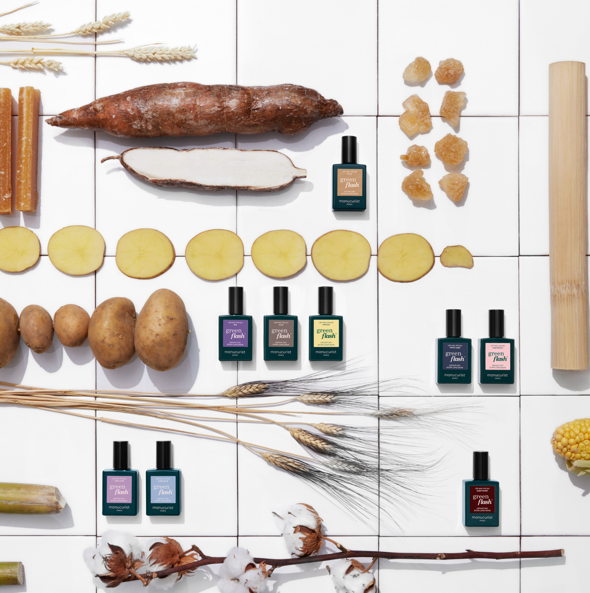 Bottles of perfume are arranged in rows on a white tiled surface, surrounded by potatoes, sliced root vegetables, wheat, corn, sugarcane, and chunks of sugar, creating a natural and colorful display.