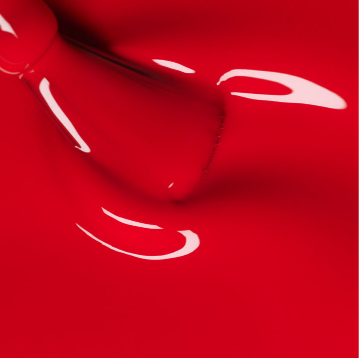Close-up of a red nail polish bottle with some polish spilled and glossy streaks, creating shiny and reflective patterns on a smooth red surface.