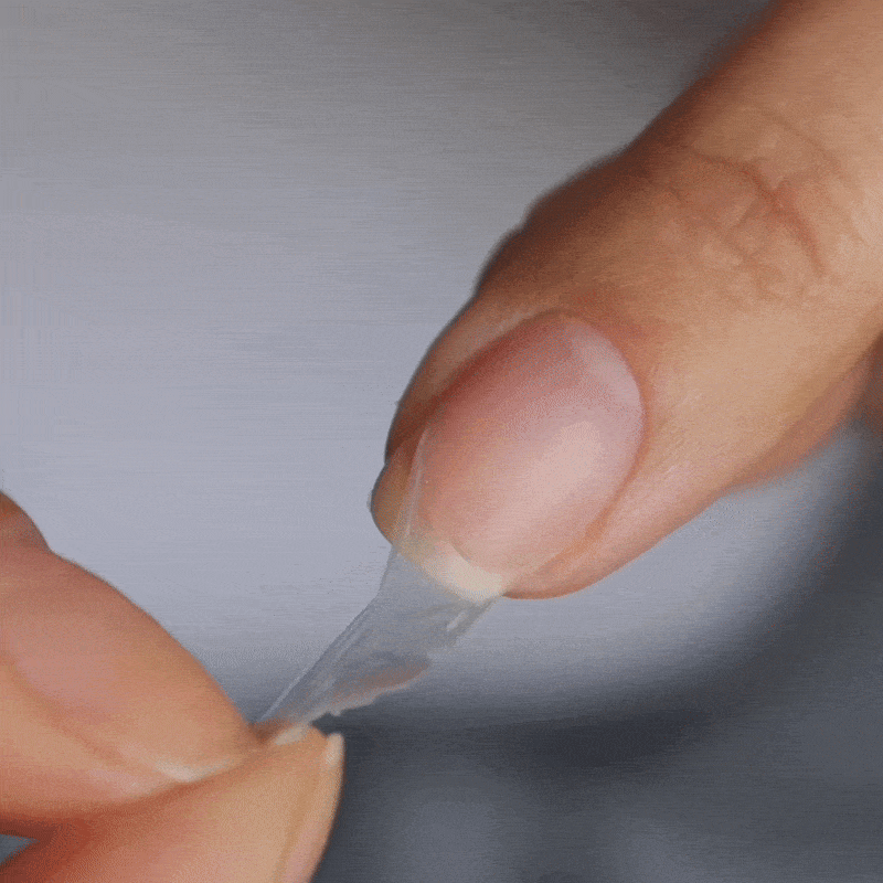 A close-up of a hand peeling a transparent adhesive film off a fingernail, revealing a clean, manicured nail underneath.