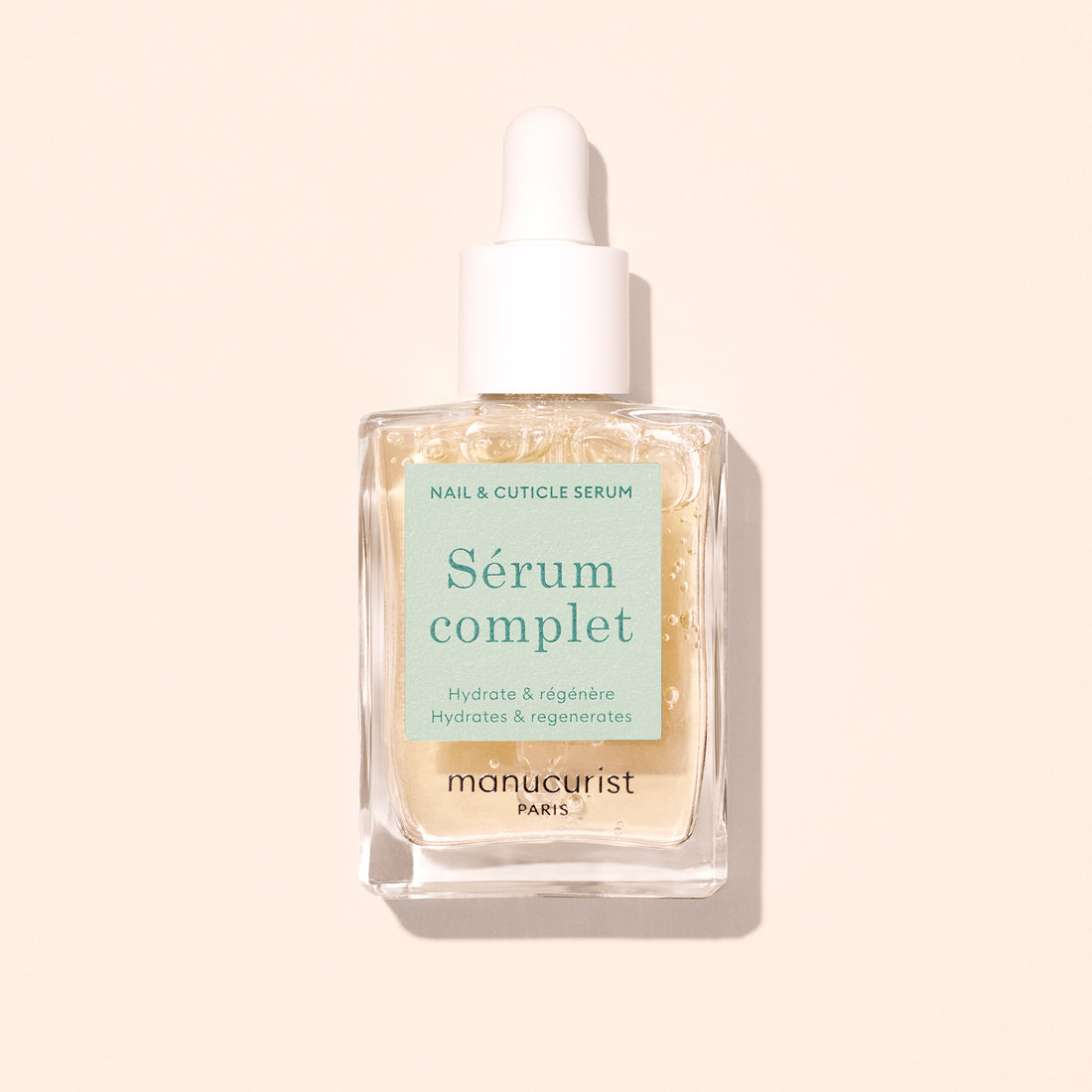 A clear glass dropper bottle labeled Nail Hydrating Serum by Manucurist, containing a nourishing nail serum with pale beige liquid inside, is shown against a light beige background.