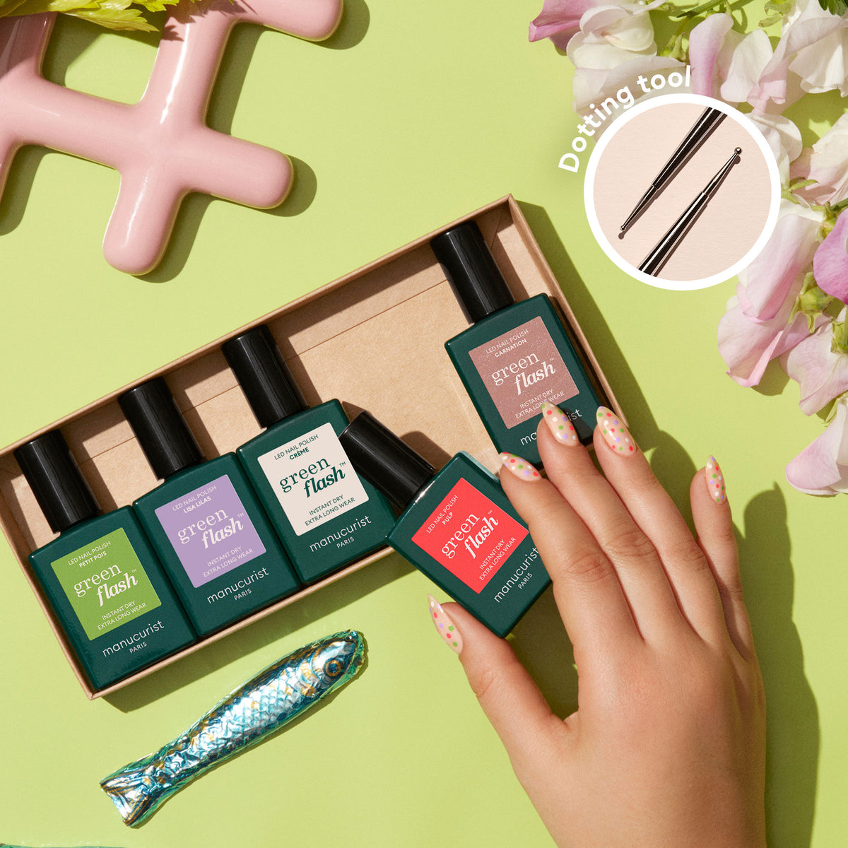 A hand with decorated nails holds a Manucurist Dot it better! near a box of five bottles, surrounded by flowers, a fish figurine, and a pink object. An inset highlights the dual-ended Dot it better! tool for creative nail art.