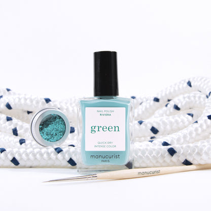 A bottle of Manucurist Underwater Glitter nail polish sits before coiled white rope with navy accents, joined by a small pot of biodegradable glitter and a fine nail art brush for creative designs.