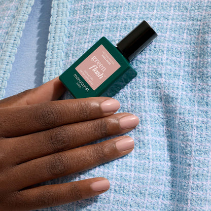 A hand with neatly manicured, Shell Beige nails holds a Manucurist Green Flash gel polish bottle against a textured light blue fabric background.