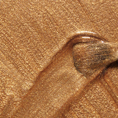 A close-up of a brush applying Manucurist’s Shimmer, spreading bronze nail polish with a honey-glazed sheen, forming a glossy streak on a golden, glittery surface.