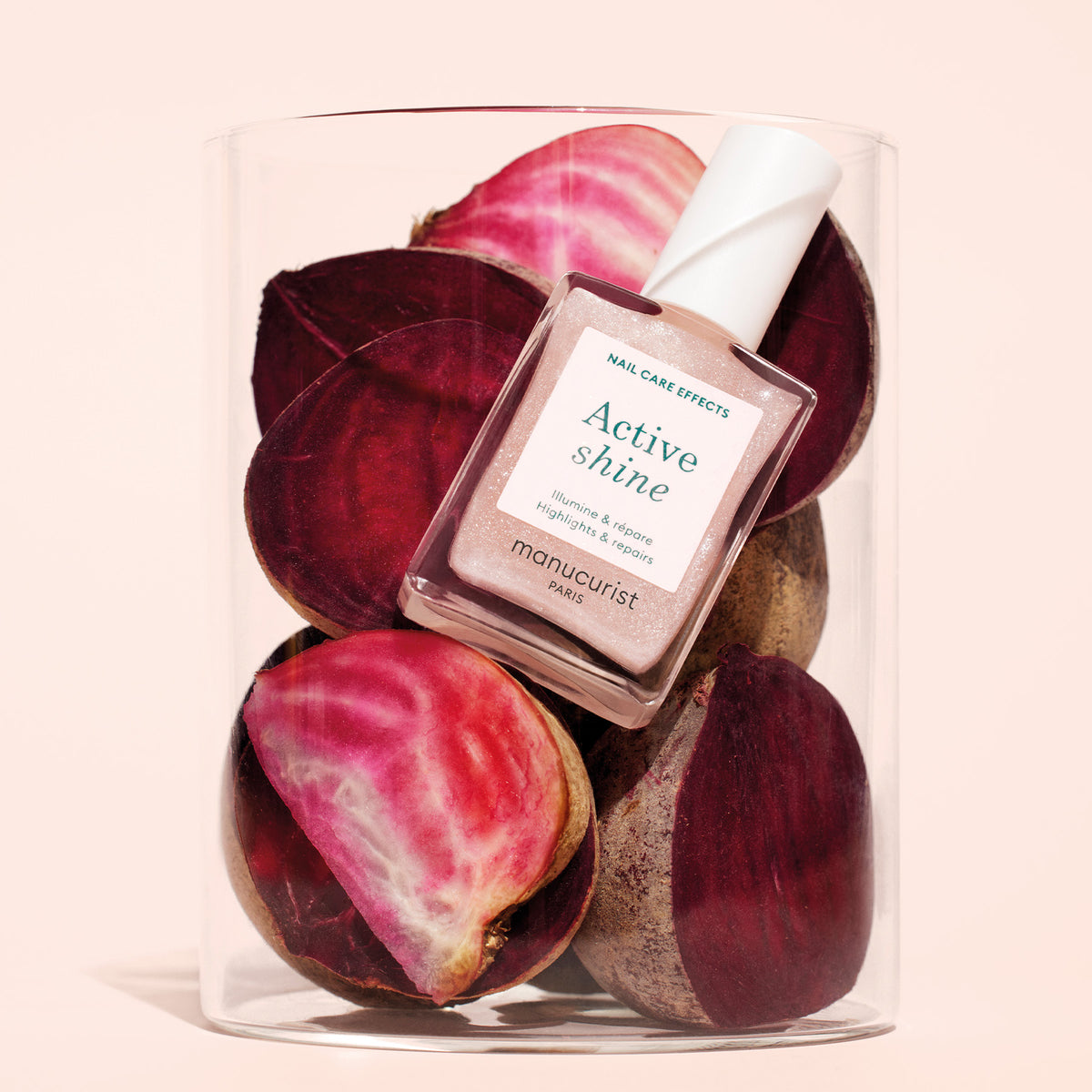 A bottle of Manucurist Active™ Shine Opal nail care rests atop sliced red and pink beets inside a clear glass container, set against a light beige background.