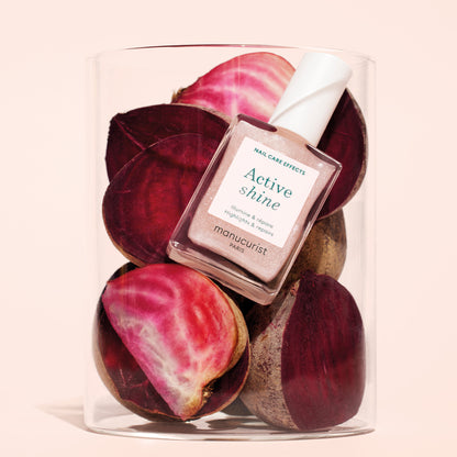 A bottle of Manucurist Active™ Shine Opal nail care rests atop sliced red and pink beets inside a clear glass container, set against a light beige background.