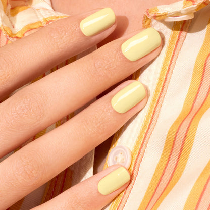 A hand with short, neatly filed nails painted in Manucurist’s Mimosa pastel yellow gel polish rests on a striped yellow, orange, and white shirt.