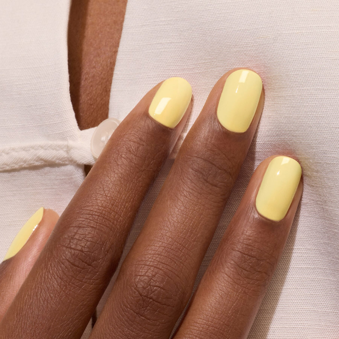 Gros plan sur une main à la peau foncée portant le vernis gel jaune pastel Mimosa de Manucurist, posée sur un vêtement blanc avec détail boutonné.