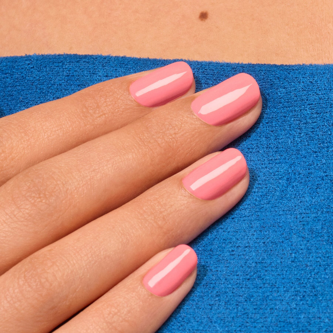 Une main aux ongles soigneusement manucurés, vernis en rose électrique Pop de Manucurist, repose sur une surface texturée bleu vif, avec un petit grain de beauté visible au-dessus.