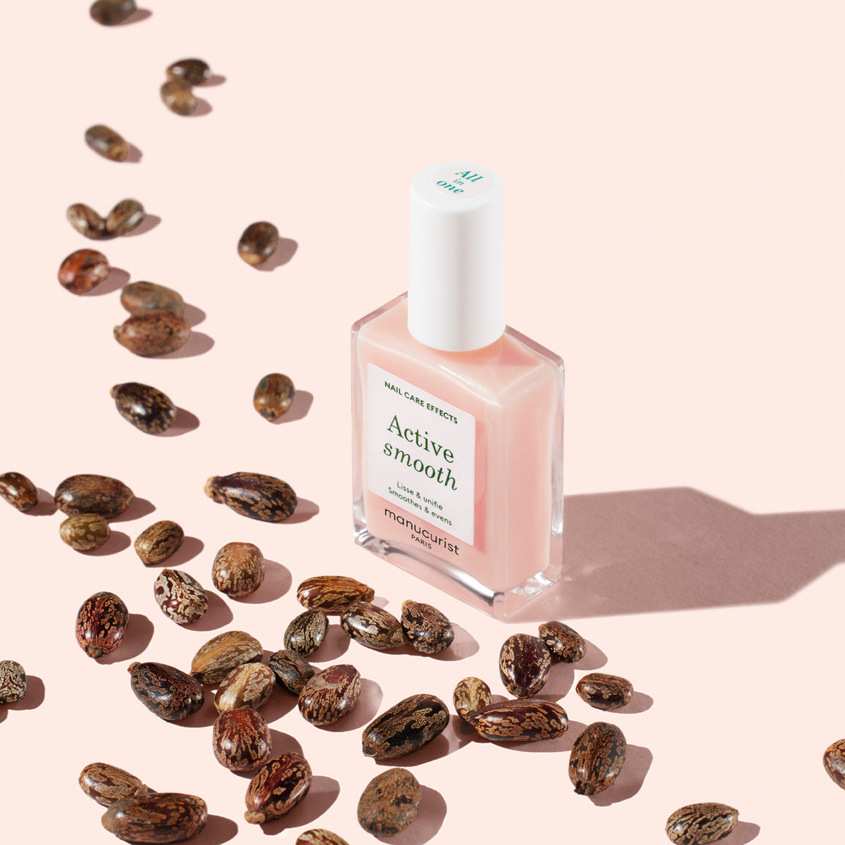 A bottle of Manucurist Active™ Smooth 00 pink nail polish sits on a light pink surface, surrounded by scattered brown seeds and casting a soft shadow—ideal for anyone seeking plant-based nails or an advanced nail treatment.