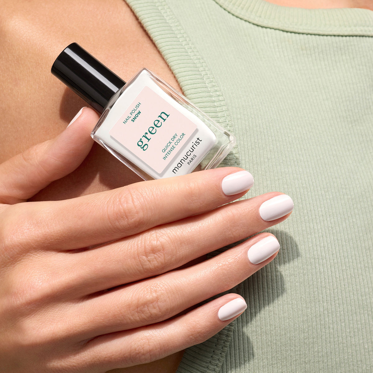 A hand with neatly manicured, pale pink nails holds a bottle of Manucurist Snow nail polish from the Green range against a sage green ribbed top. The label on the bottle reads green by Manucurist.