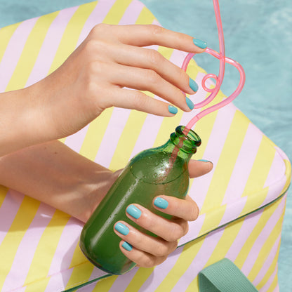 Wearing Manucurist’s Riviera, a person with turquoise blue nails holds a green glass bottle with a curly pink straw, hand resting on a yellow and white striped cushion by an energizing blue pool.